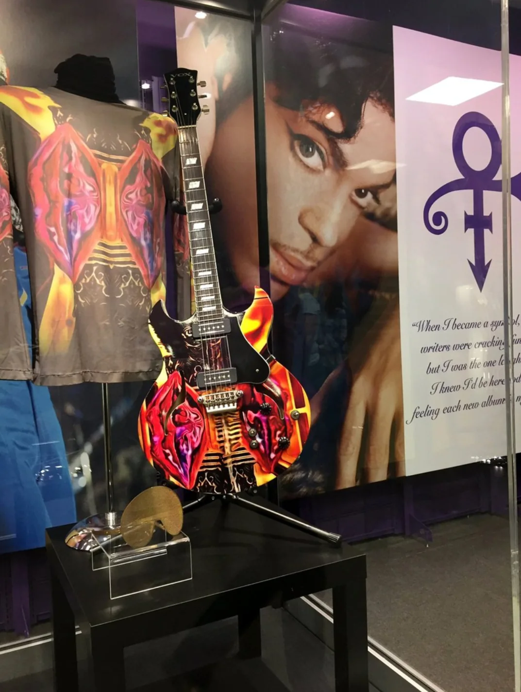 Graphic-wrapped guitar on a stand in a glass display beside a matching garment and a portrait of Prince