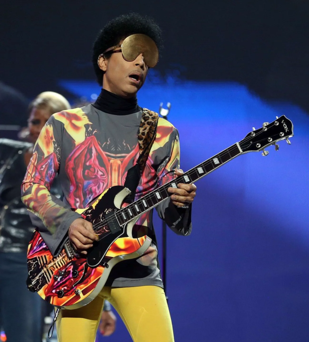 Prince on stage holding a guitar with red, yellow, and black graphics against a blue-lit background