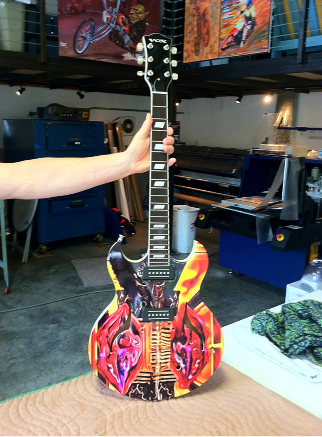 Red, yellow, and black graphic-wrapped guitar held upright over a padded workbench in the workshop